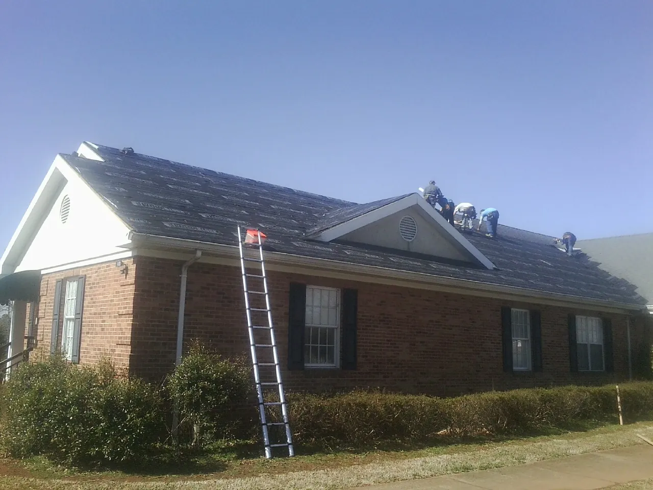 Crew tearing shingles off a brick ranch roof with a ladder set — Emergency Roof Repair work by our crew in Elbow Lake