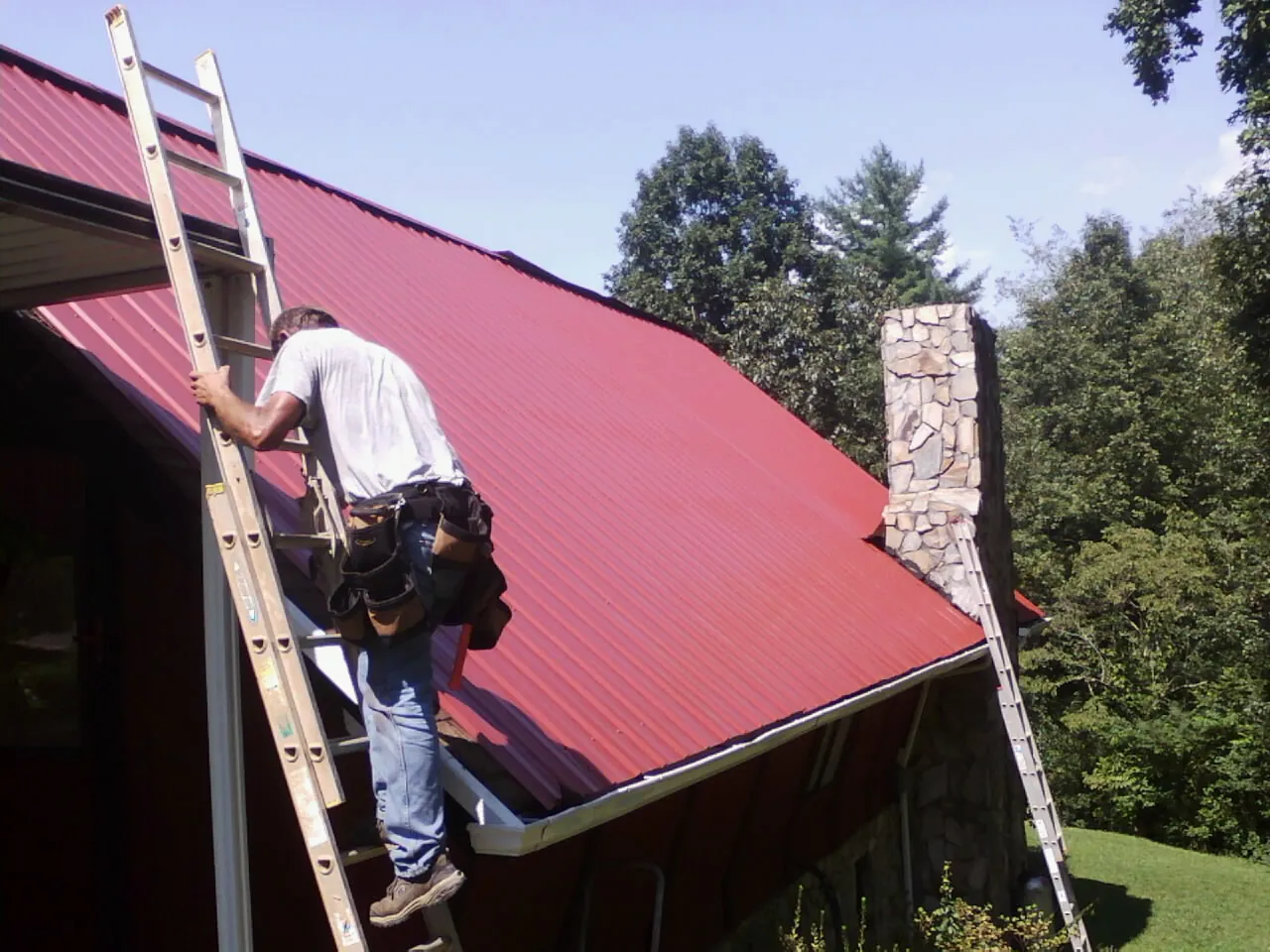 Roofer on a ladder beside a red metal roof and stone chimney — Soffit & Fascia Repair by Roofing Brothers in Elbow Lake